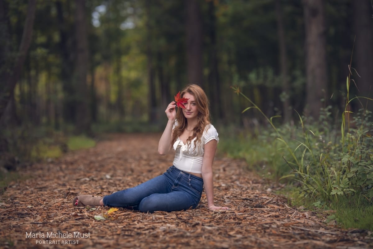 Detroit-area High school senior picture photographer captures a graduation photo of a girl resting on on a bed of leaves as she holds up one leaf to obstruct her eye - taken in a Michigan forest
