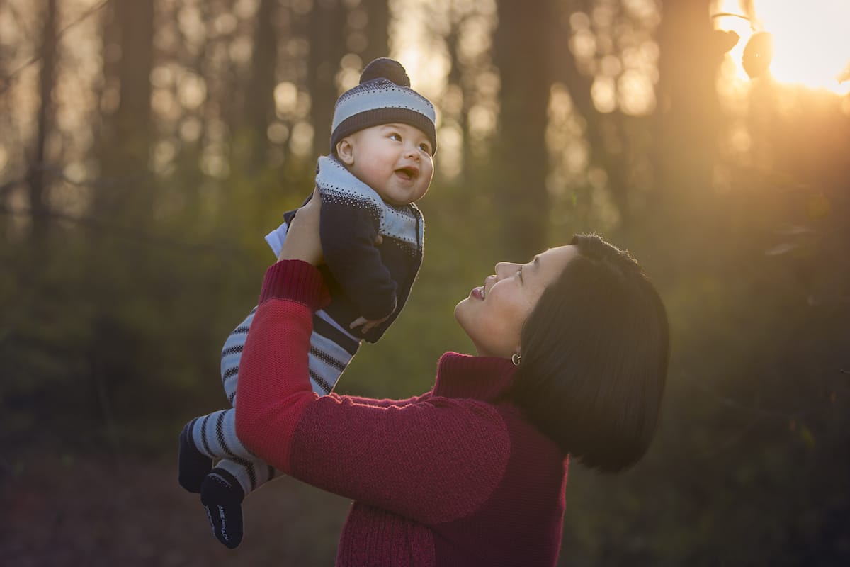local family photographer takes photo of a Detroit mother and daughter smiling as the mother lifts the toddler in the air, a fall background behind them