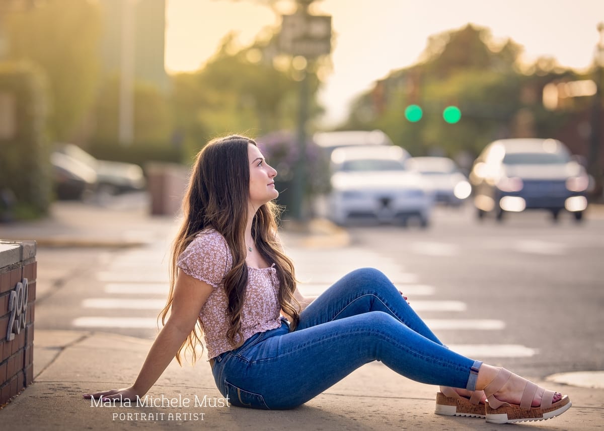 Detroit-area High school senior picture photographer captures a graduation photo of a girl smiling as she sits at a profile view of the camera - a busy downtown Detroit metro area is behind her.
