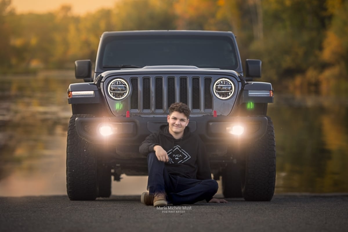 Detroit-area High school senior picture photographer captures a graduation photo of a boy sitting in front of his jeep and smiling at the camera