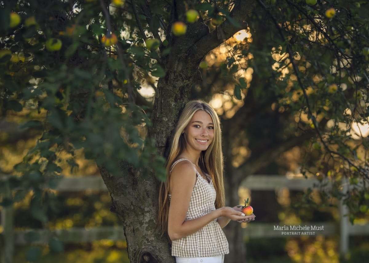 Detroit-area High school senior picture photographer captures a graduation photo of a girl leaning against a tree at sunset and smiling at the camera