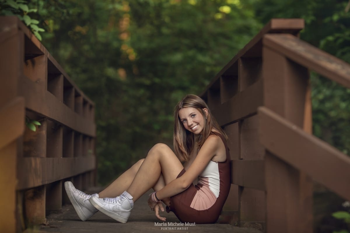 Detroit-area High school senior picture photographer captures a graduation photo of a girl smiling at the camera as she holds her knees and sits along the edge of a Michigan park bridge