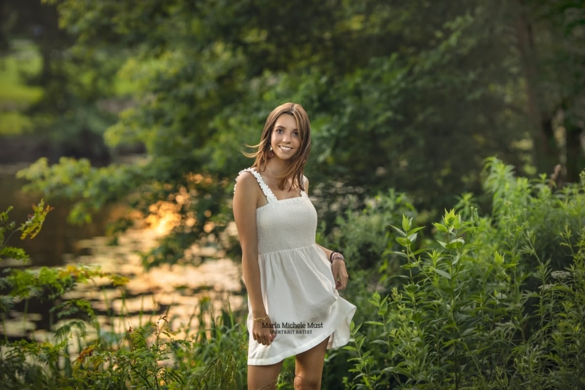 Detroit-area High school senior picture photographer captures a graduation photo of a girl in a flowing dress posing in a forested area lit by the sunset