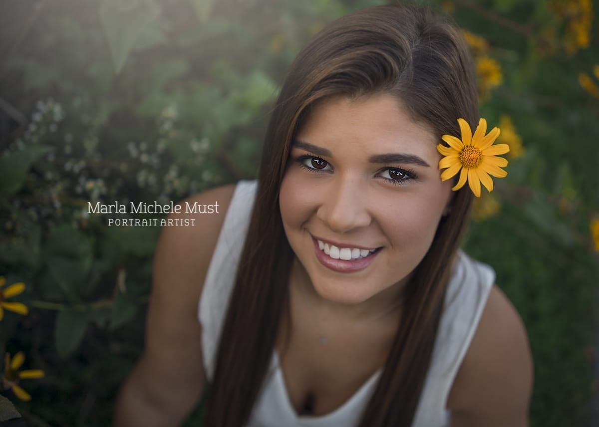 Detroit-area High school senior picture photographer captures a graduation photo of a girl smiling up at the camera with a flower in her hair
