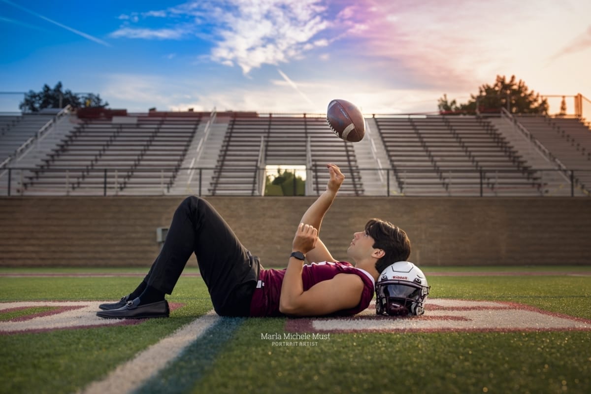 Detroit-area High school senior photographer captures the portrait of aboy laying on his back and throwing a football in a Detroit high school football field