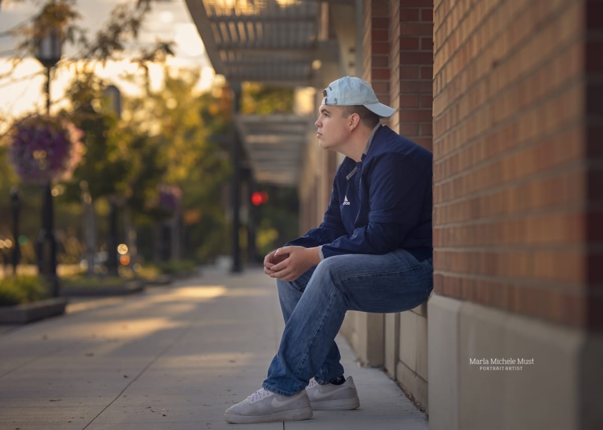 High school senior poses for his senior portrait while looking into the distance and sitting on the ledge of a building in a Detroit-area metro