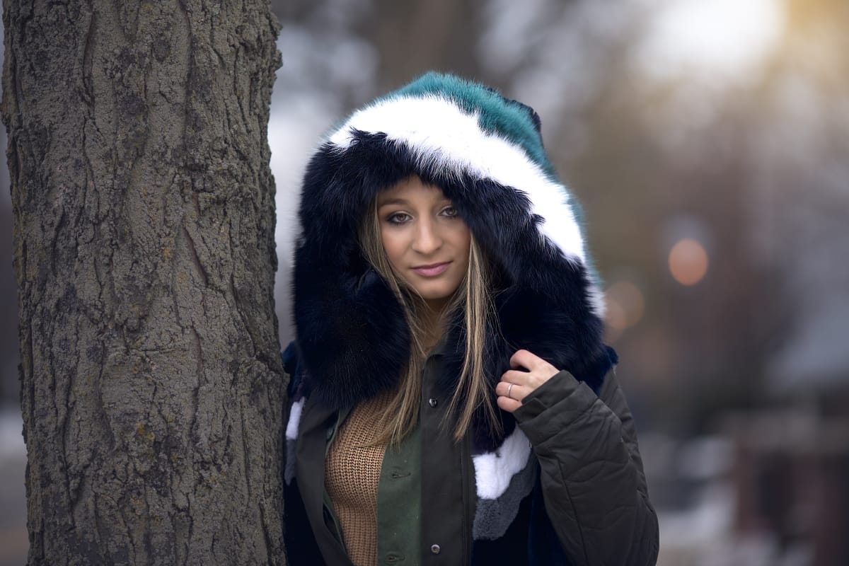 High school senior poses in a big, fluffy winter coat with the hood pulled up in a Detroit-area park