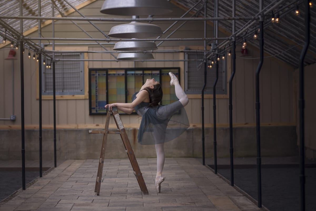 Ballerina practices leg extensions while leaning against a ladder in a dancer portrait taken in a warehouse with cozy string lights around her.