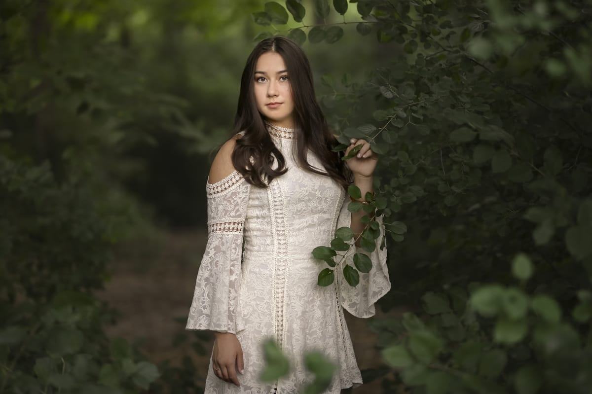 Girl poses for her senior portrait in a fantasy-inspired dress among a foreground of Detroit area hedges and leaves