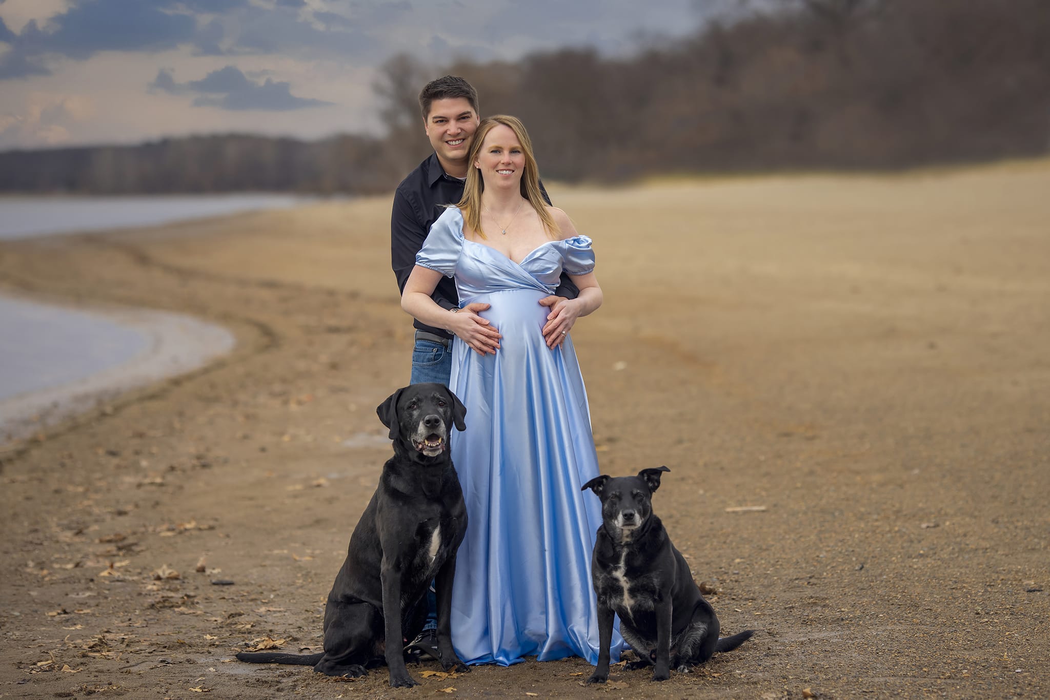 A pregnant woman, her partner and their two dogs smile and stand on a Michigan beach for a maternity photoshoot; a moment captured by a skilled maternity photographer.