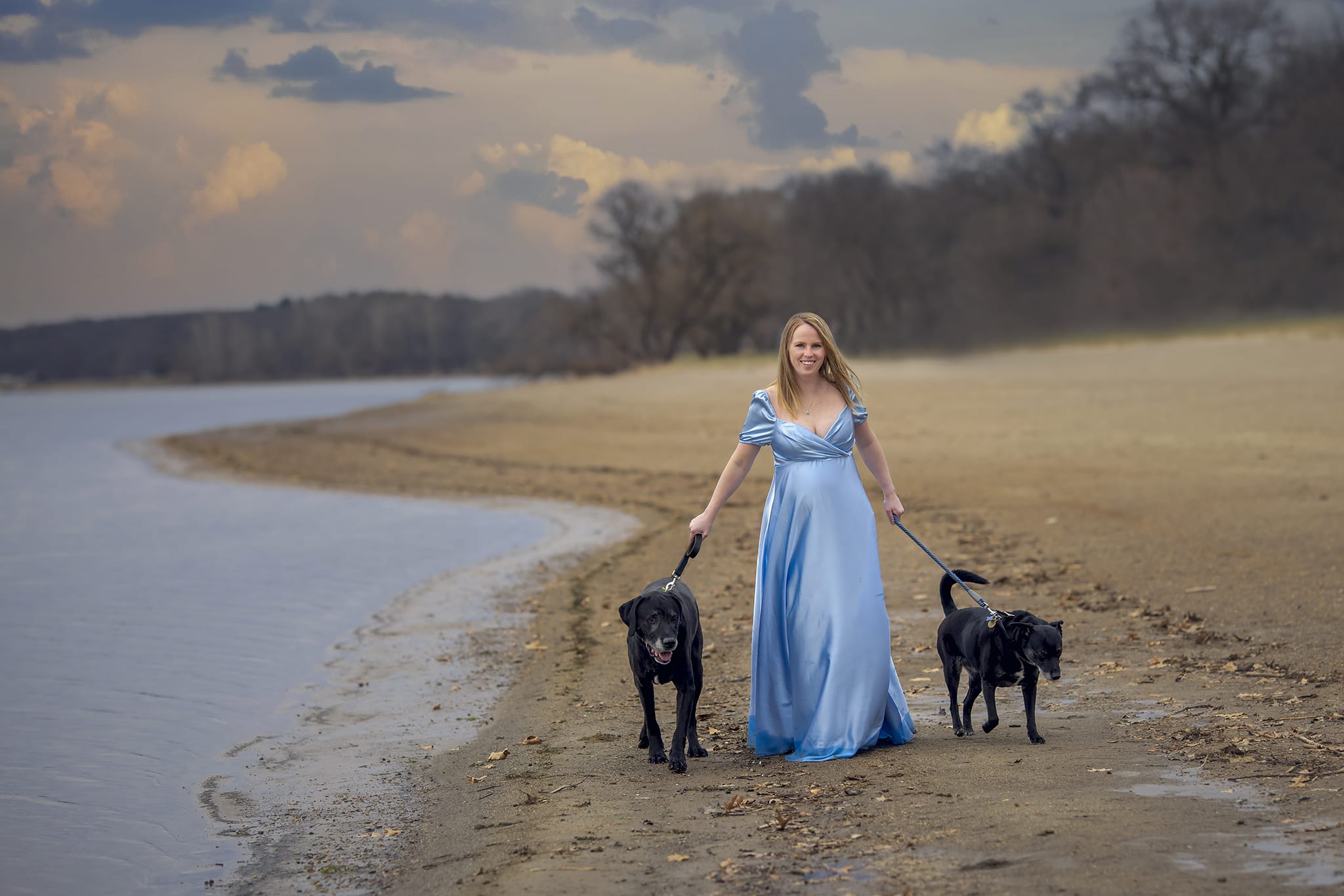 A pregnant woman in her two dogs smile and walk along a Michigan beach for a maternity photoshoot; a moment captured by a skilled maternity photographer.