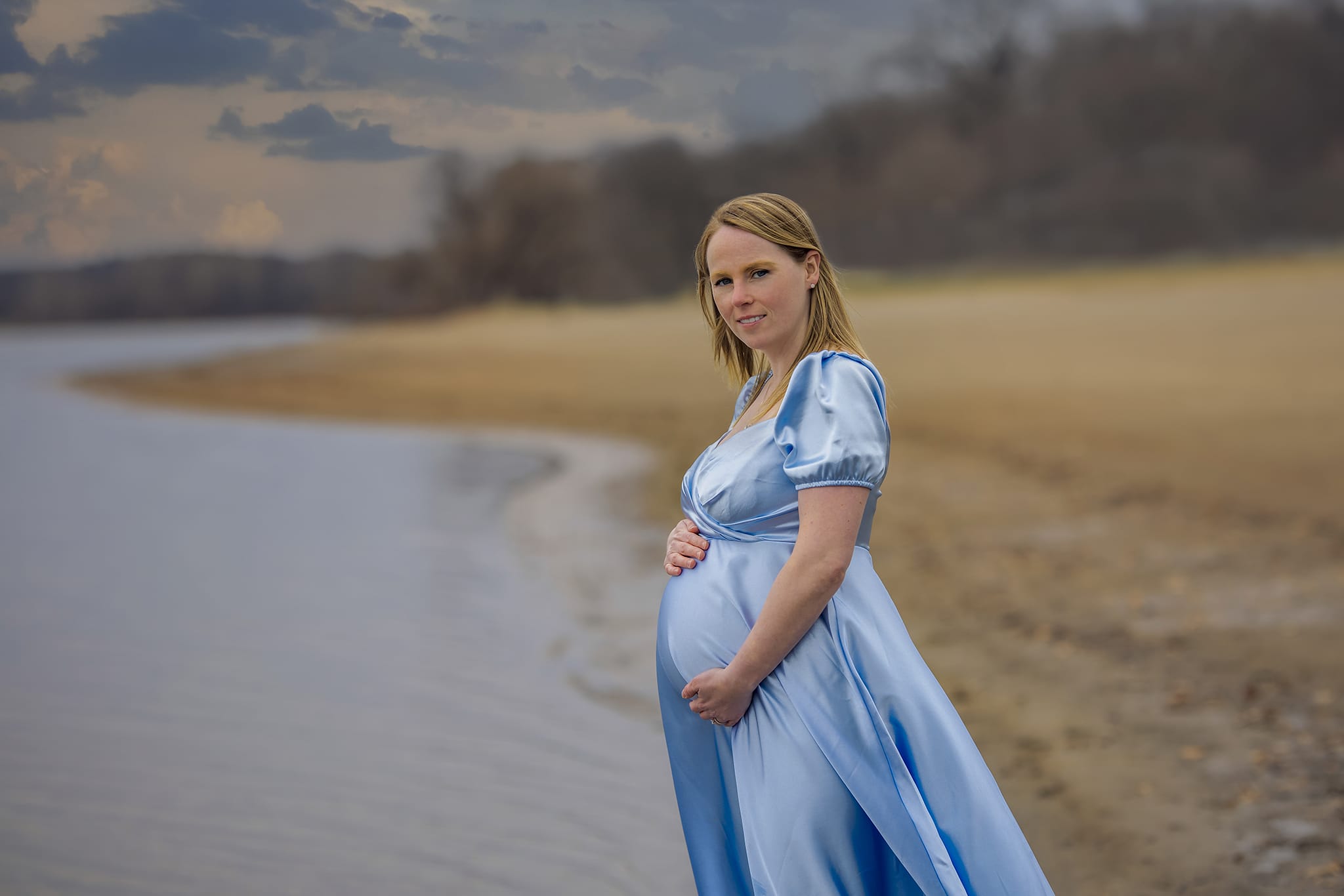A pregnant woman in a dress gracefully holds her belly as she stands on a beach for a Detroit-area maternity photoshoot.