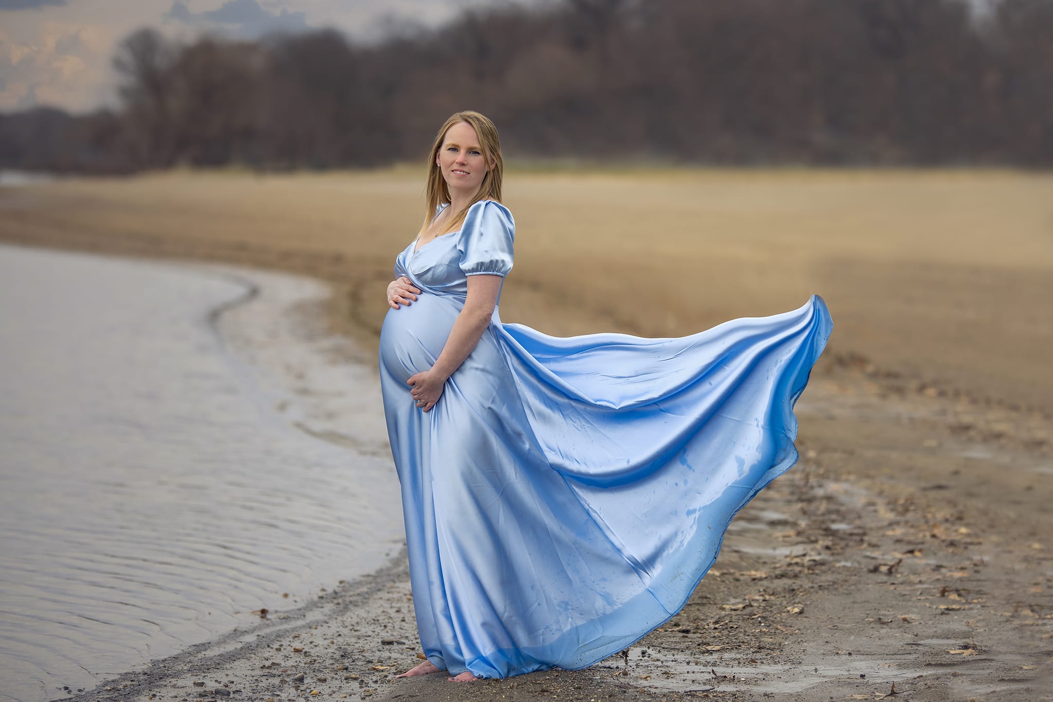 A pregnant woman in a dress gracefully holds her belly as she stands on a beach for a Detroit-area maternity photoshoot.