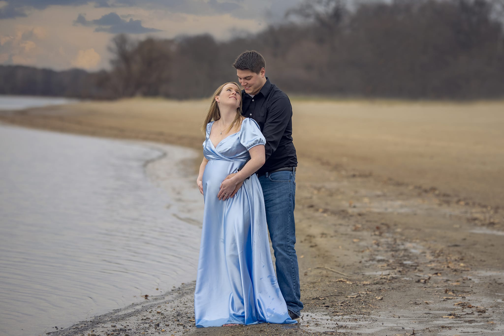 A pregnant woman stands in front of her partner as her partner holds her from behind on a Michigan beach for a maternity photoshoot; a moment captured by a skilled maternity photographer.
