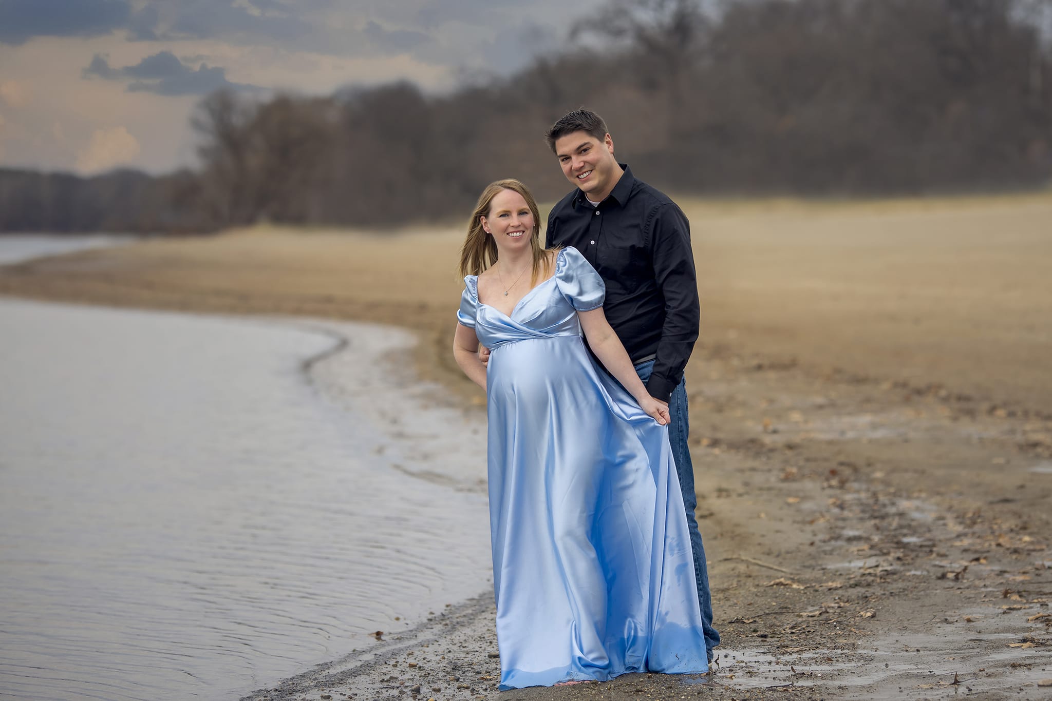 A pregnant woman stands in front of her partner as her partner holds her from behind on a Michigan beach for a maternity photoshoot; a moment captured by a skilled maternity photographer.