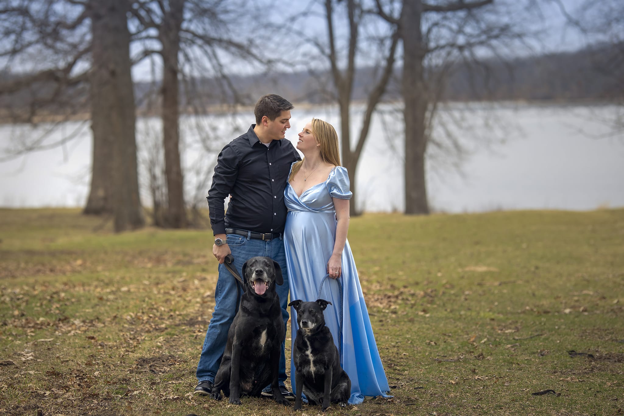 A pregnant woman, her partner, and their two dogs stand in a forest near a Michigan beach for a maternity photoshoot; a moment captured by a skilled maternity photographer.