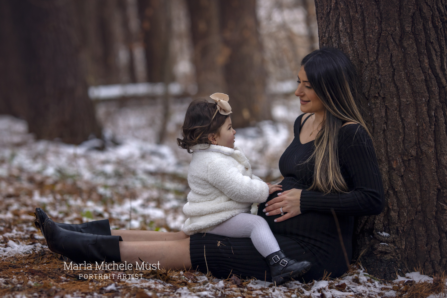 An expecting mother smiles at her child sitting on her lap in a snowy Detroit forest; a moment captured by a Michigan maternity photographer.