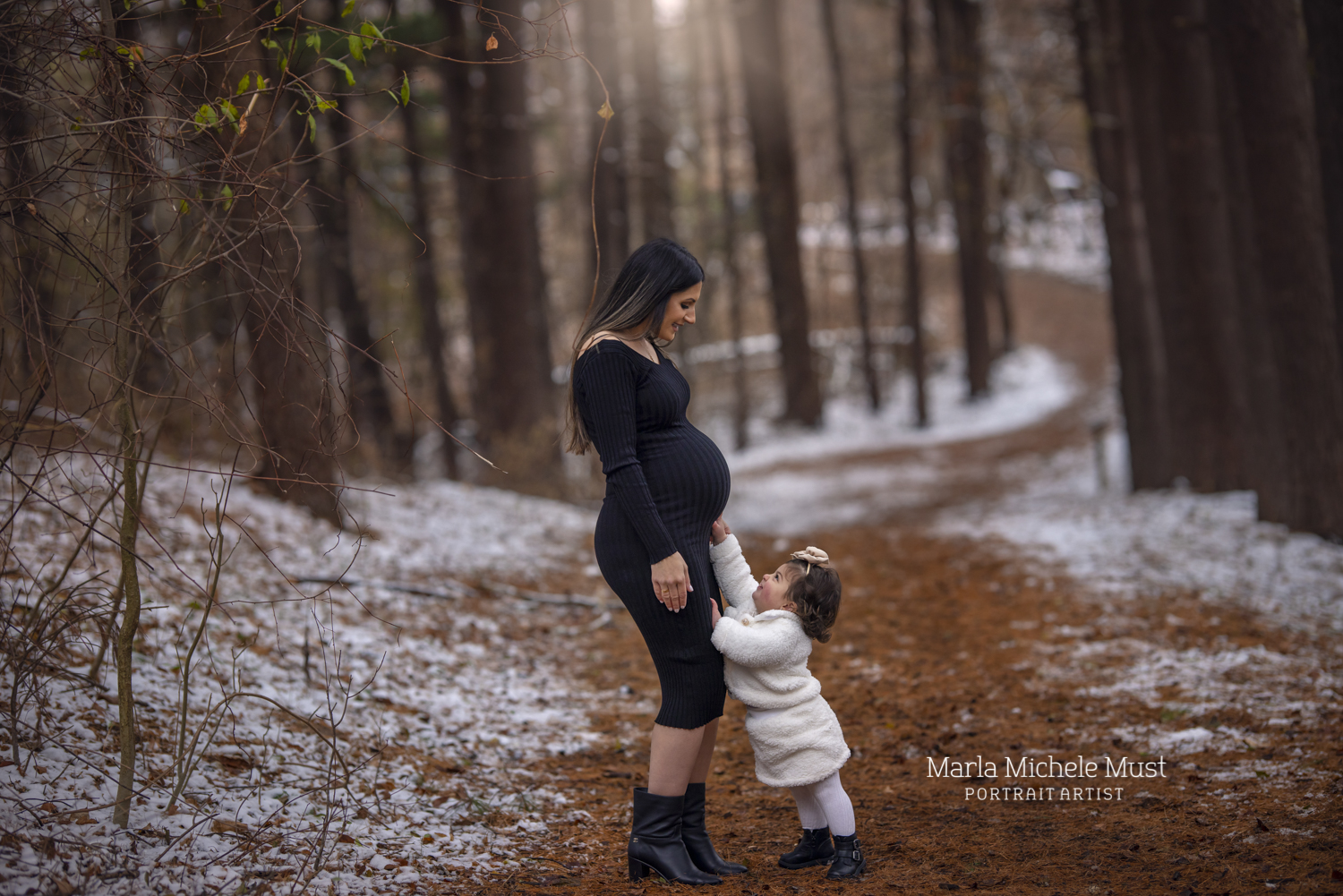 An expecting mother smiles at her young daughter as she reaches towards her pregnant belly in a snowy Detroit forest.
