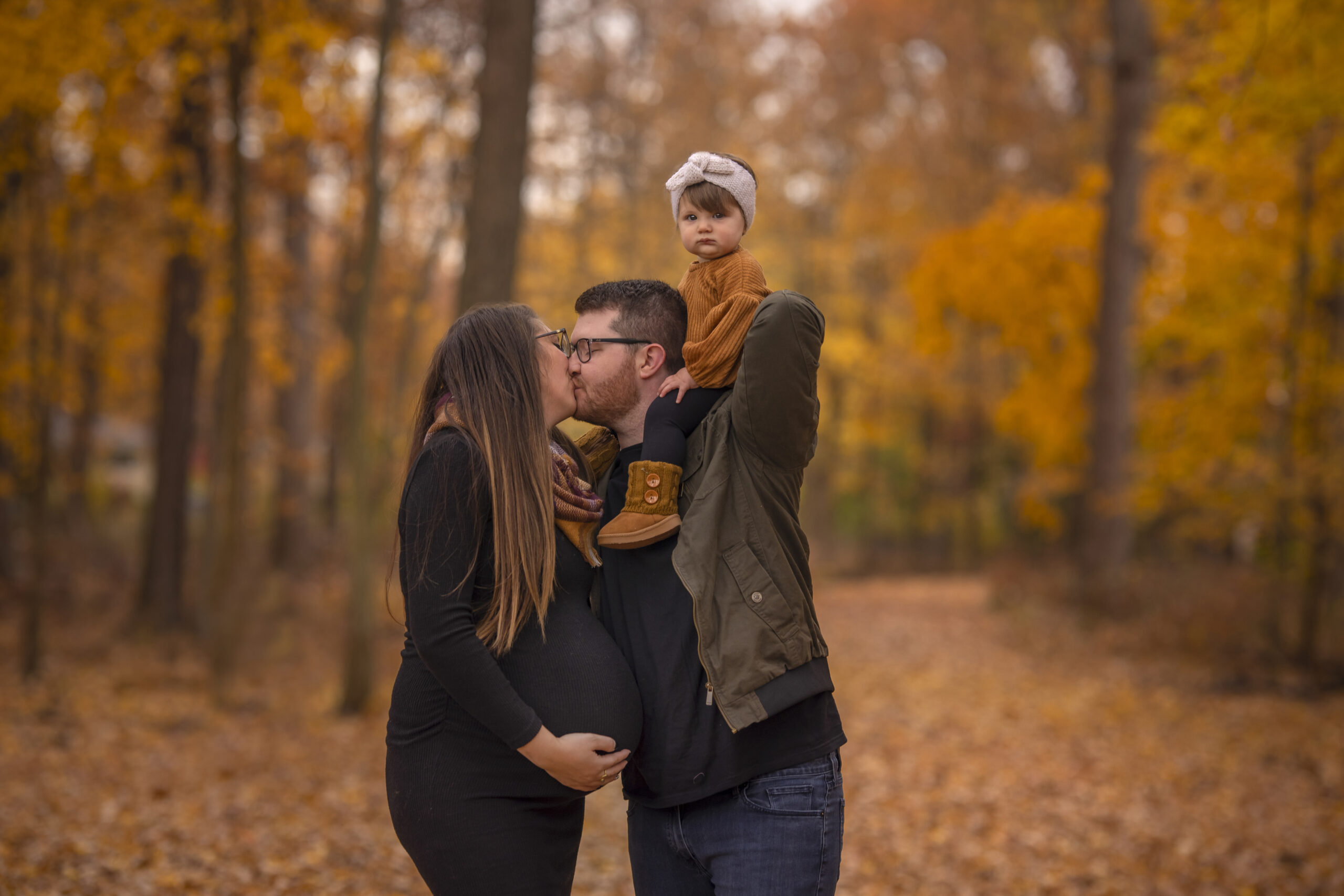 A couple kiss during a maternity photoshoot in Detroit. They are standing in a forest with their oldest child on the husbands shoulders.