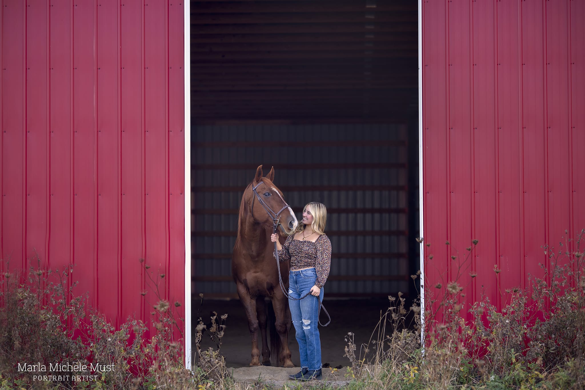 A rustic photograph by a Detroit equine photographer, featuring the owner standing alongside their horse in the doorway of a large red barn.