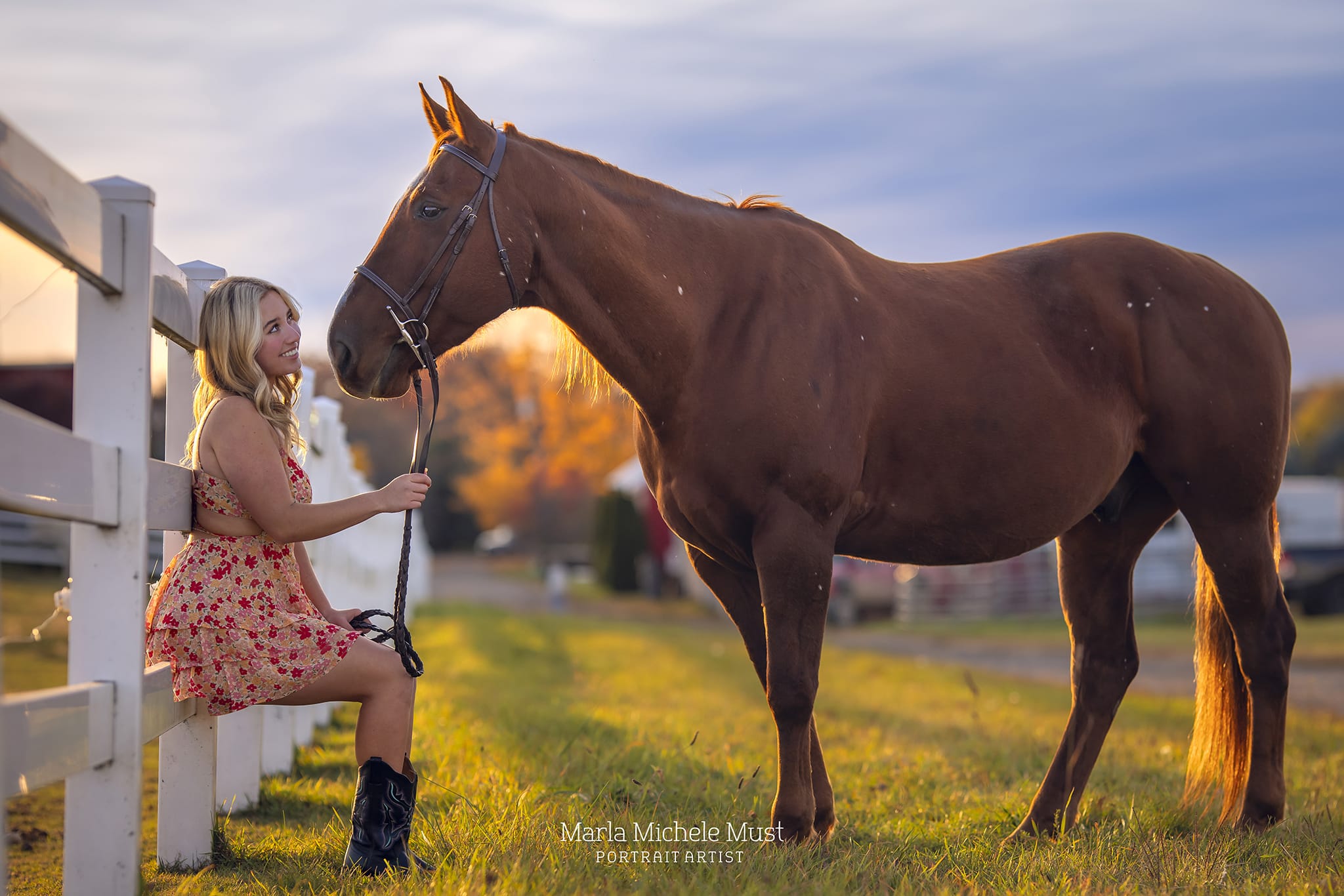 Detroit equine photographer's captivating shot: Woman gently holding reins and looking up at her horse while sitting on a white fence during golden hour..