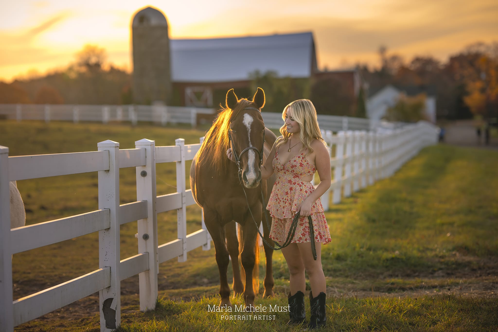 A captivating equine moment captured by a Detroit photographer, featuring a woman standing alongside a horse during sunset, a red barn and fence in the background.