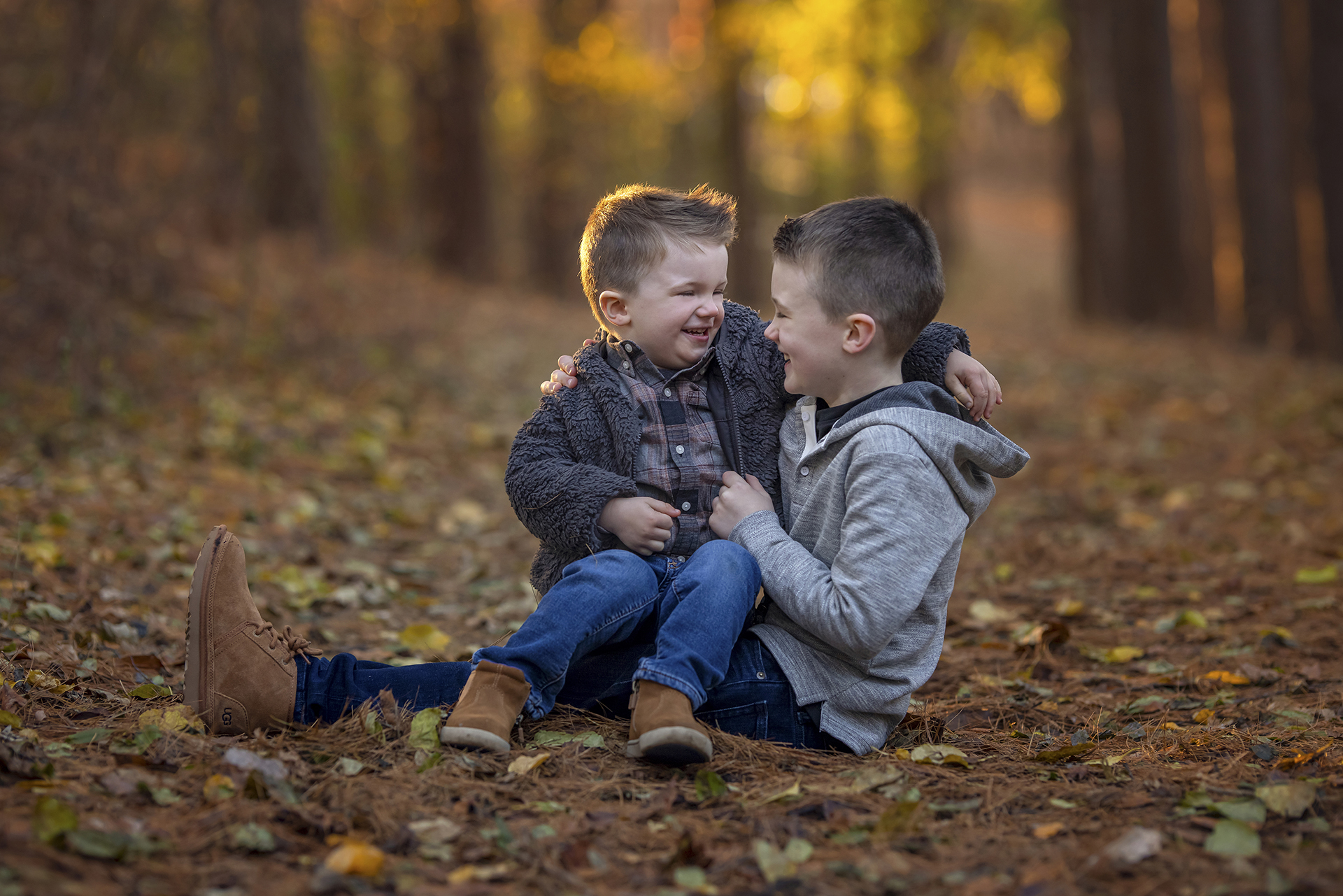 Two young sibling boys sit with each other among autumn leaves and laugh during a Detroit photoshoot.