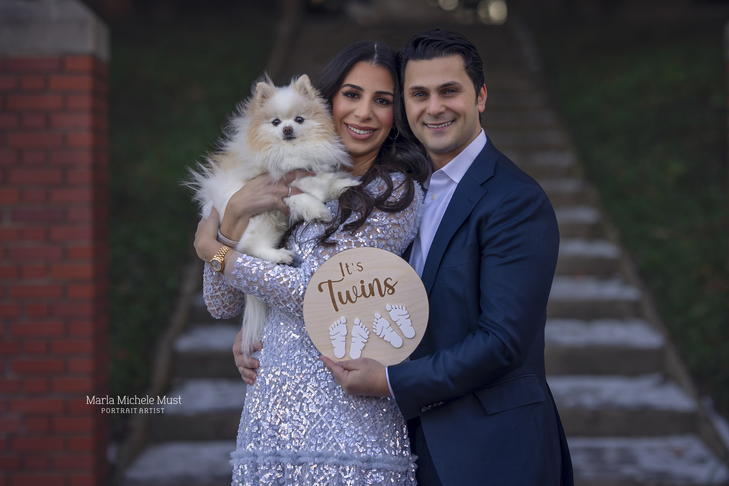 A formally dressed expecting couple (a small dog in their arms) hold a small, round, wooden sign displaying the words "It's Twins!" with two pairs baby footprints imprinted upon the surface; a moment captured by a Detroit maternity photographer.