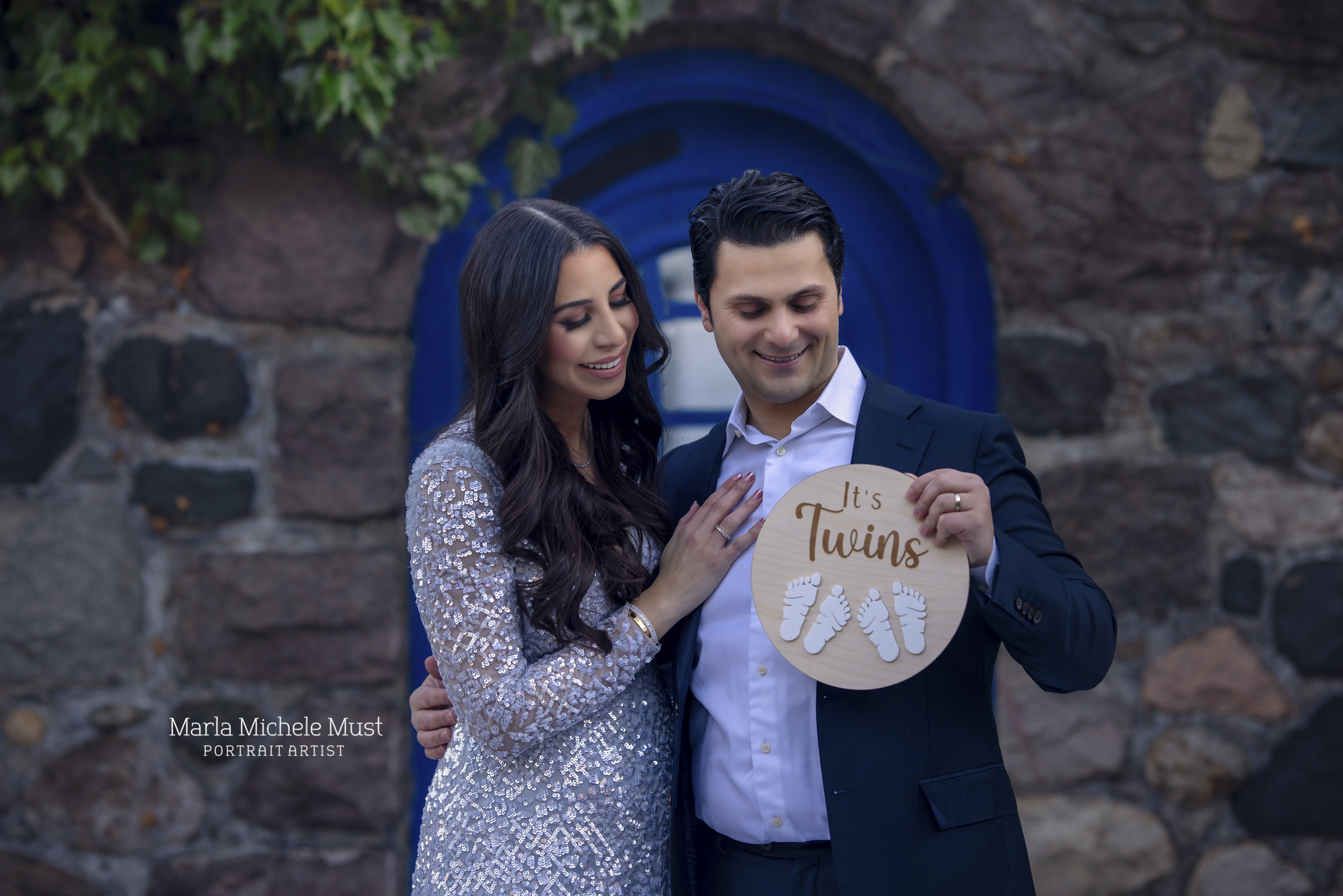 An expecting couple, formally dressed, hold a small, round, wooden sign displaying the words "It's Twins!" with two pairs baby footprints imprinted upon the surface; a moment captured by a Detroit maternity photographer.