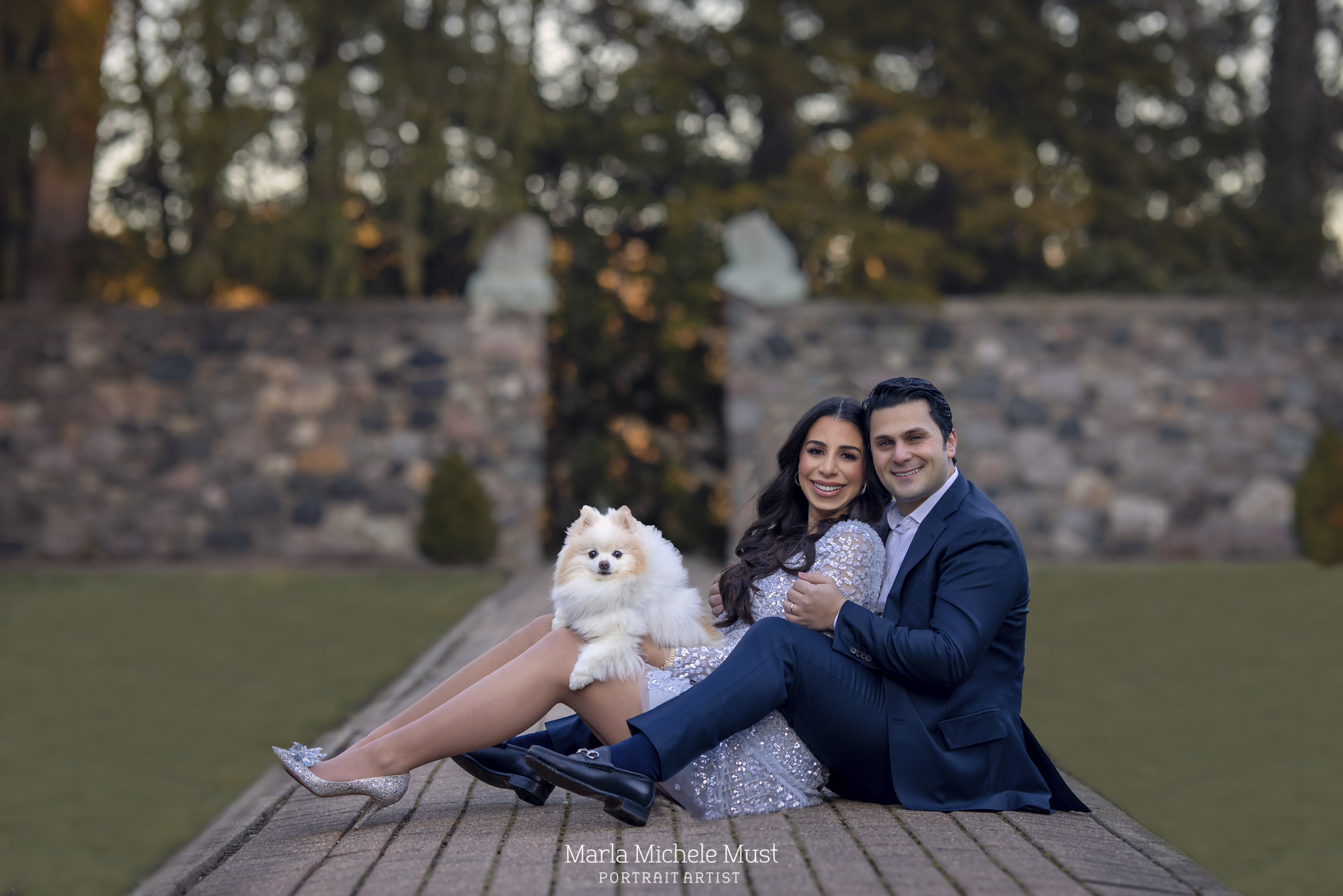 A formally dressed expecting mother, her partner, and their small dog on their lap, sit gracefully on a pathway in a Detroit park for a maternity photoshoot; a moment captured by a talented Michigan maternity photographer.