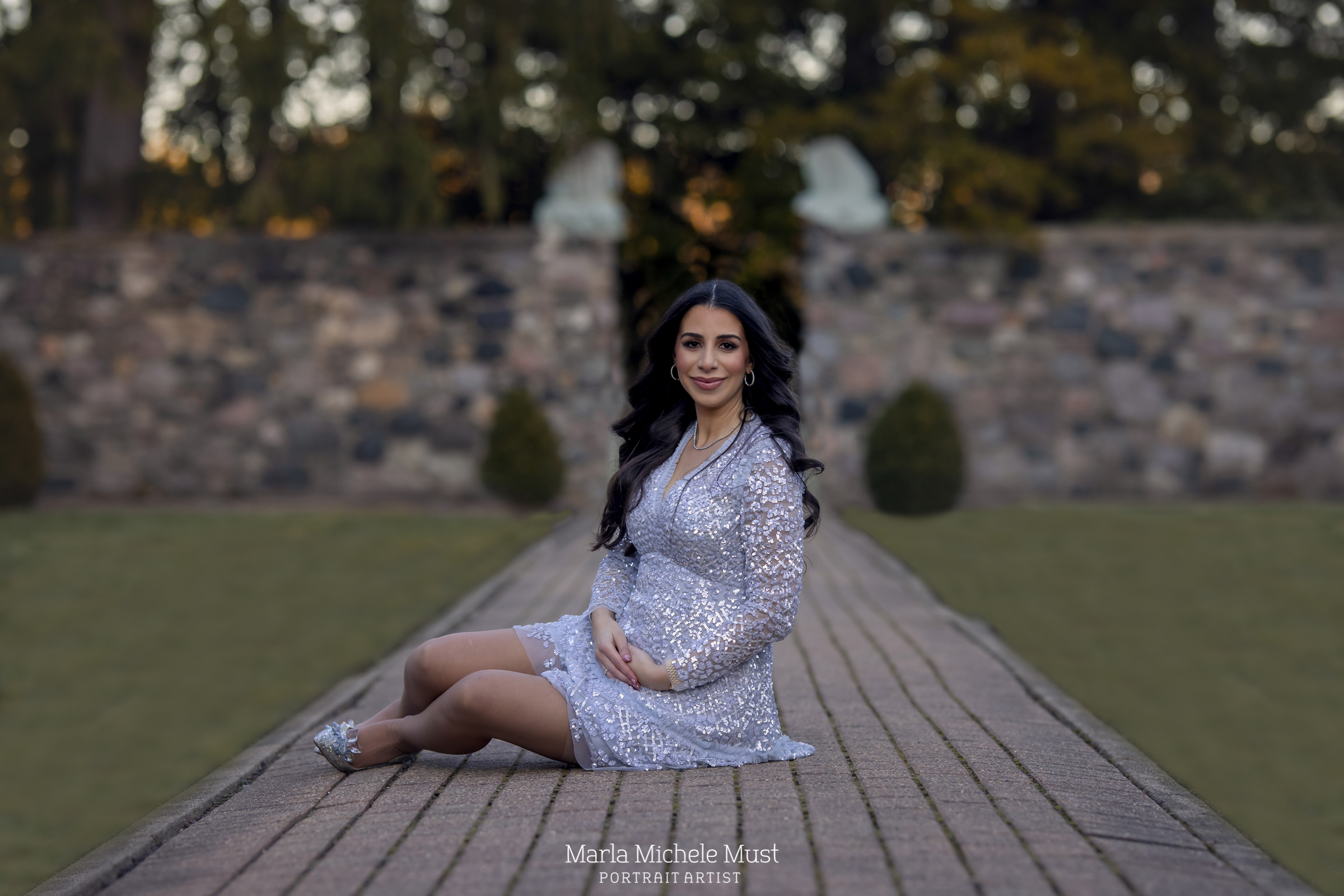 A formally dressed expecting mother sits gracefully on a pathway in a Detroit park for a maternity photoshoot; a moment captured by a talented Michigan maternity photographer.