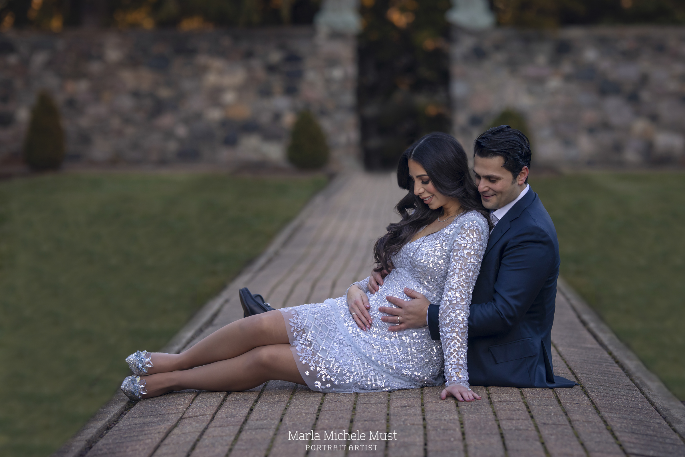 A formally dressed expecting mother and her partner sit together on a pathway in a Detroit park for a maternity photoshoot; a moment captured by a talented Michigan maternity photographer.