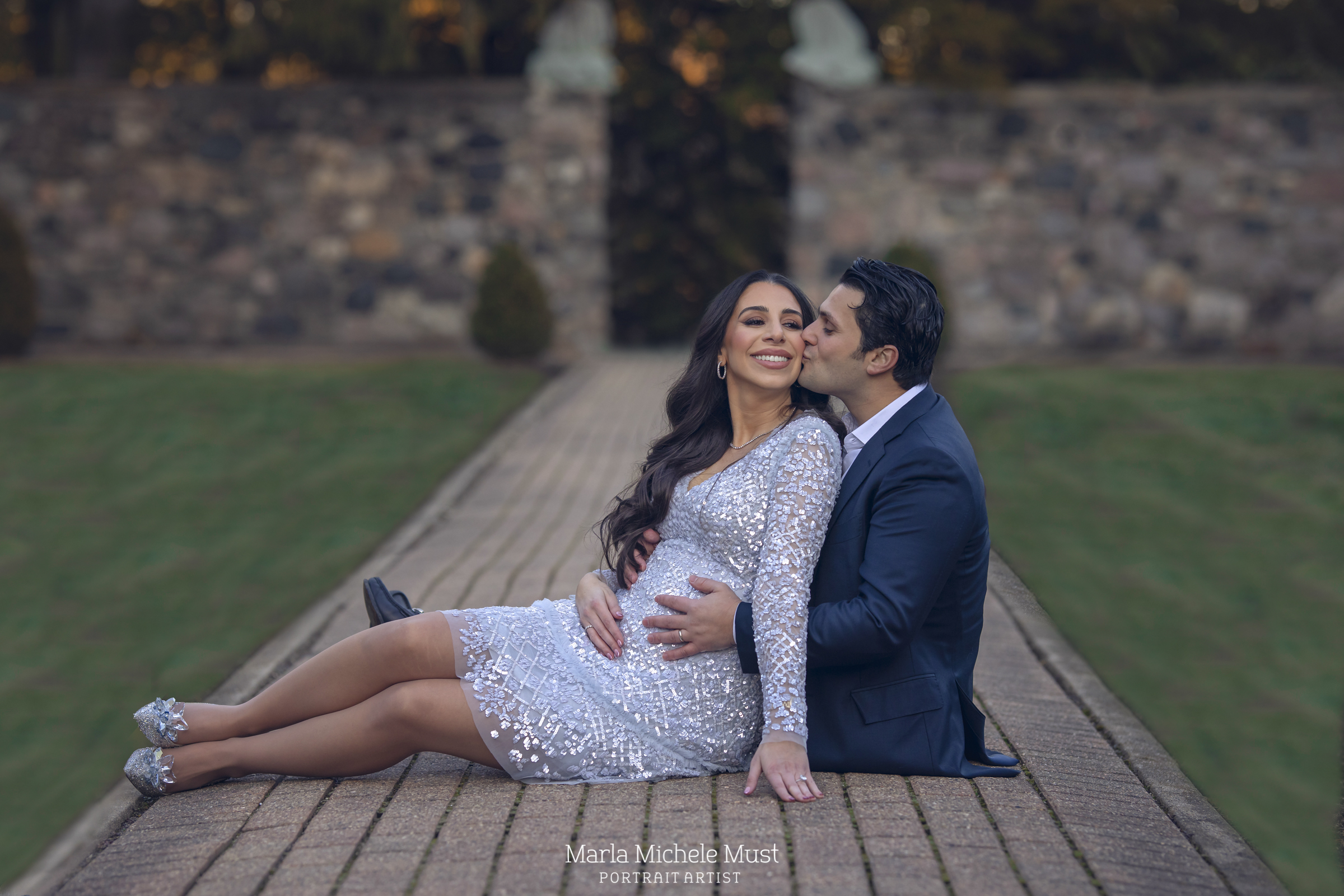 A formally dressed expecting mother and her partner sit together on a pathway in a Detroit park for a maternity photoshoot; a moment captured by a talented Michigan maternity photographer. .