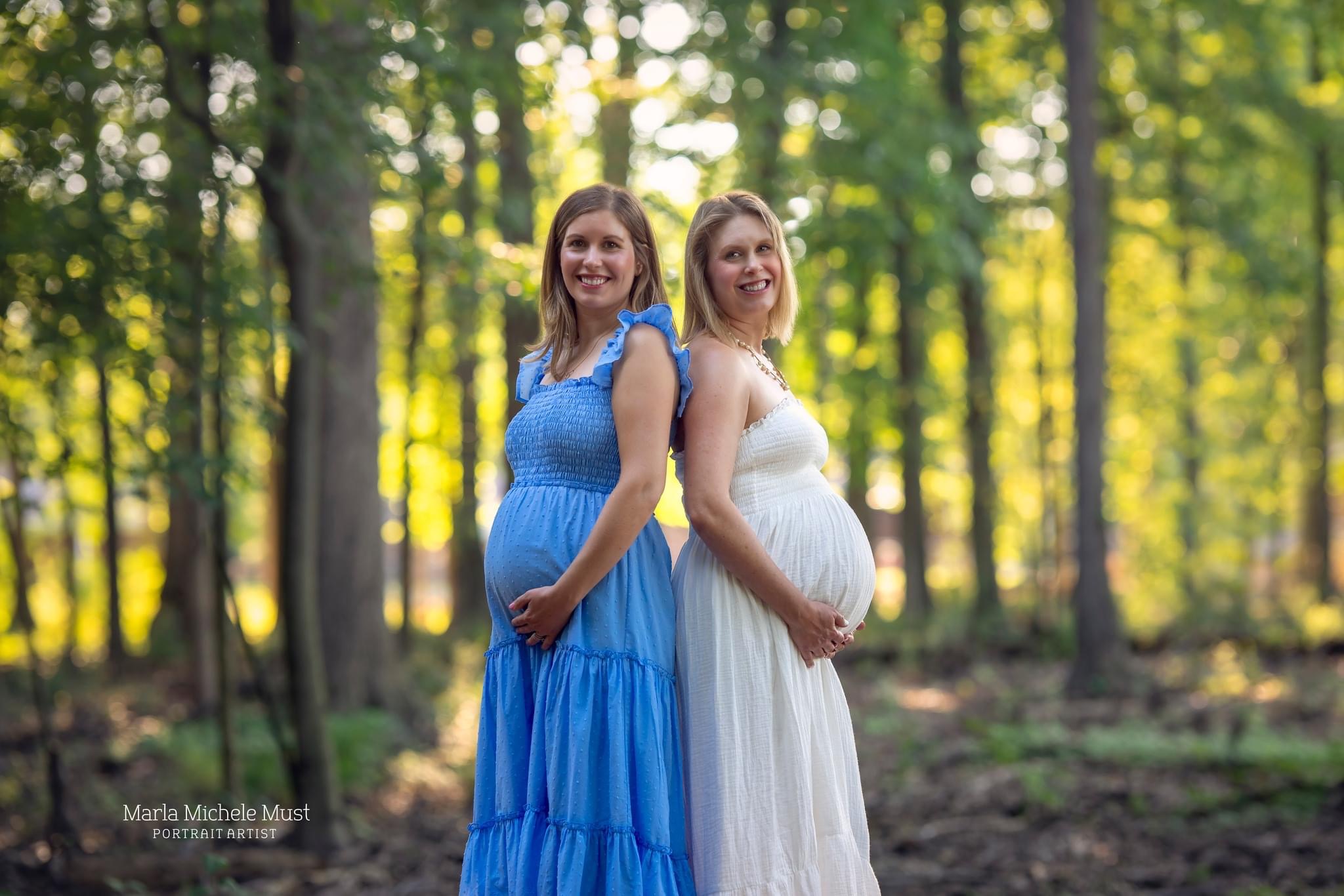 Two women in long, flowy dressed stand back to back in a forest while holding their pregnant bellies; a moment captured by a Detroit maternity photographer.