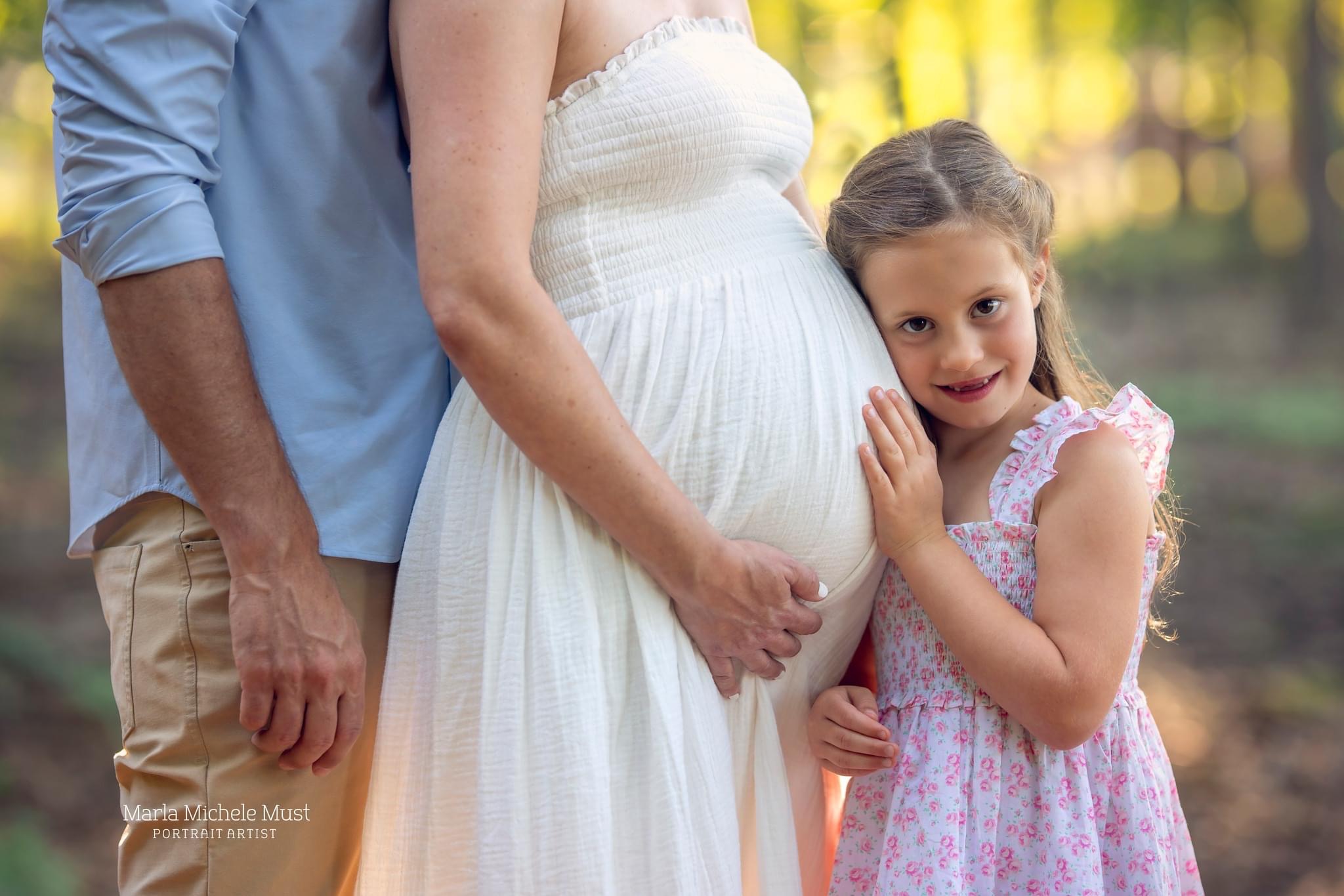 Close up shot of a father standing behind a pregnant mother, and their young daughter leans her head on her mother's pregnant belly,; a moment lovingly captured by a Detroit maternity photographer.