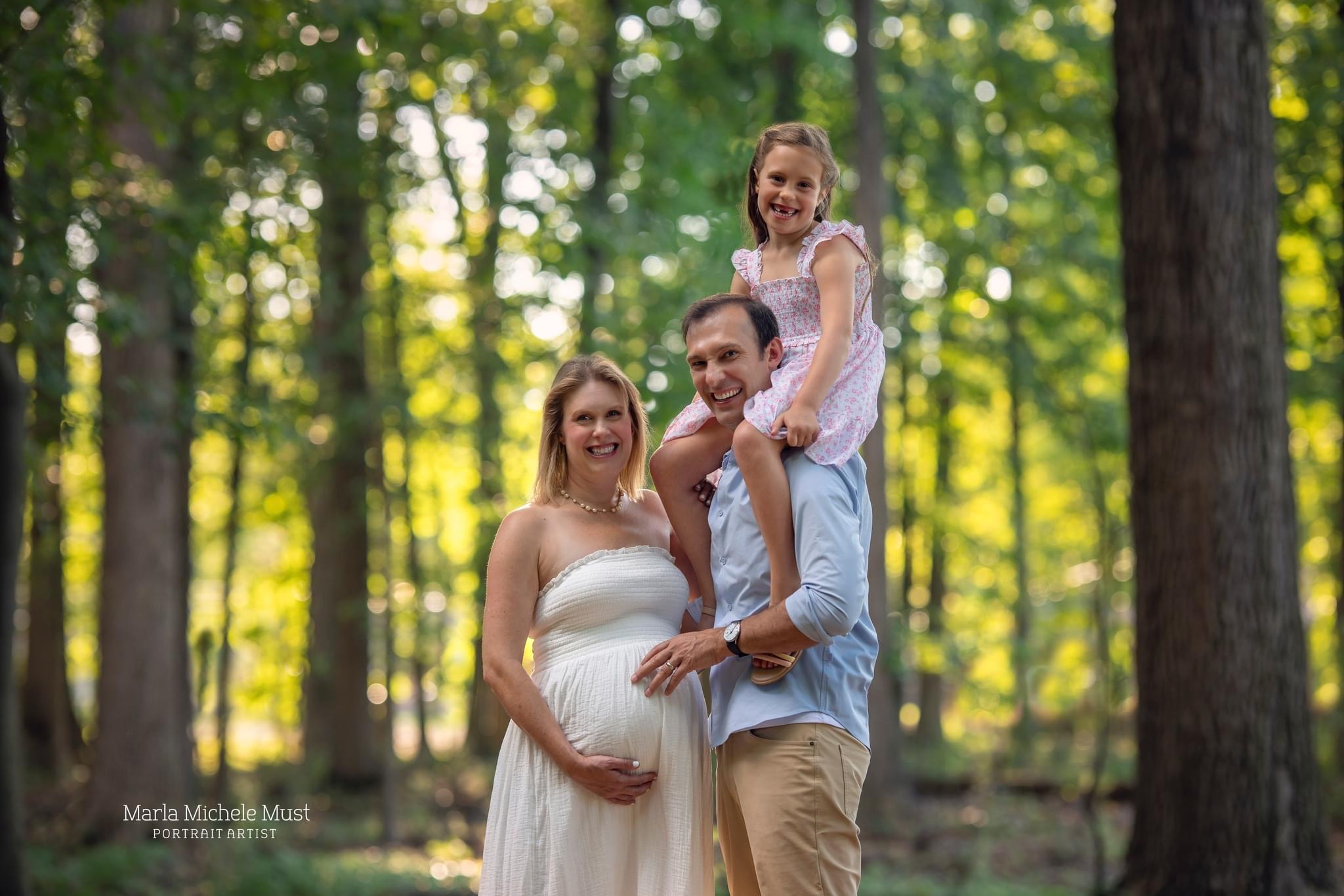During maternity photoshoot, an expecting mother stands in a Michigan forest with her partner, their daughter is sitting on his shoulders.