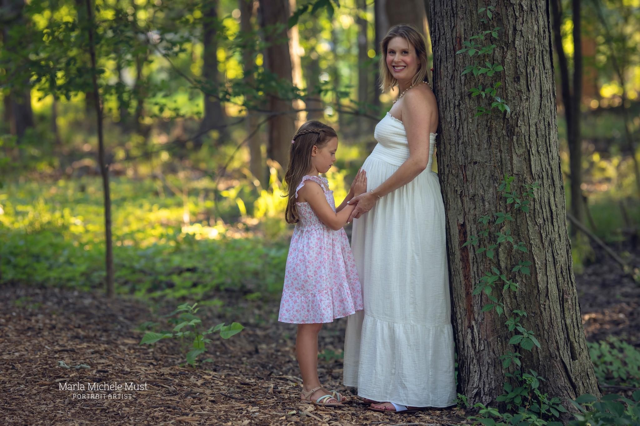 An expecting woman stands against a tree in a Detroit, her daughter lovingly placing a hand on her belly; a moment captured by a Michigan maternity photographer.