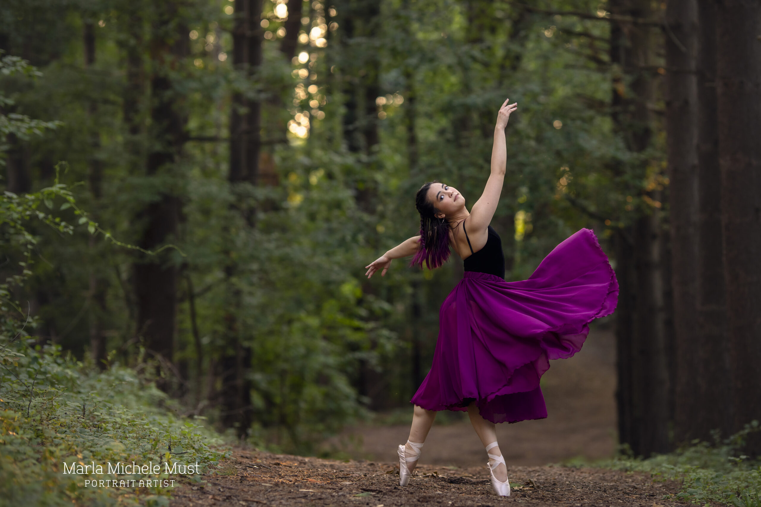 Dancer extends her arms upwards as she poses on a forest pathway during a dance photoshoot, taken by a Michigan dance photographer.